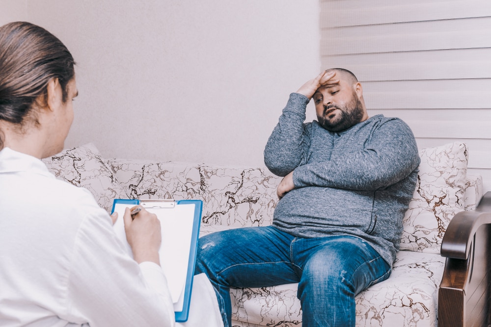 Un hombre con obesidad se ve preocupado mientras habla con una profesional de atención de salud.