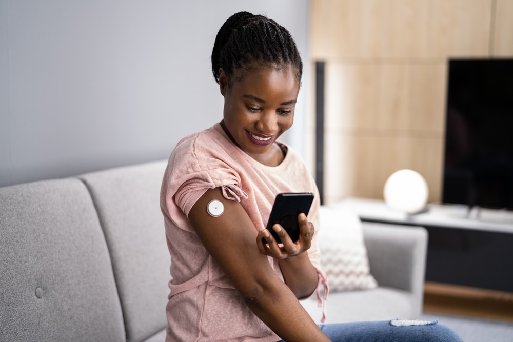 A woman sits on a couch and checks her blood glucose level with a CGM and mobile phone.