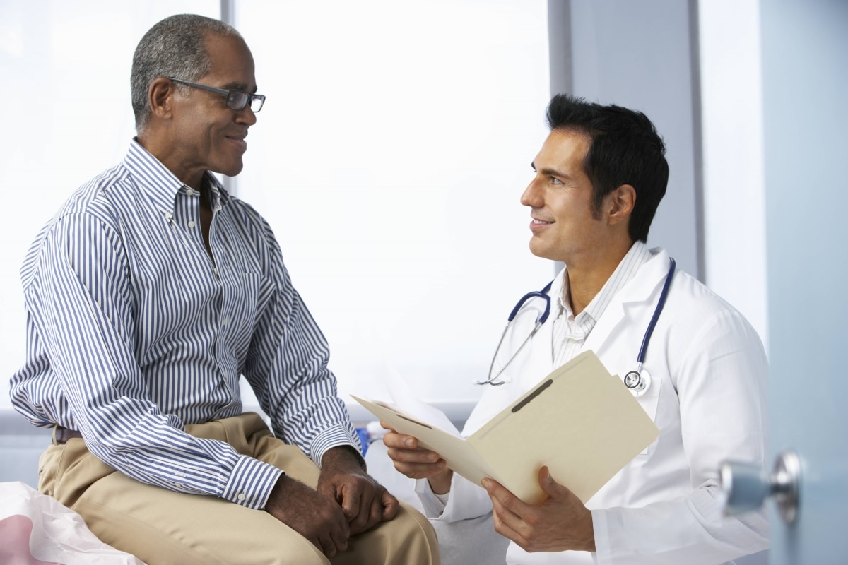 A male patient talks with a male health care professional while seated on an exam table in a medical office.