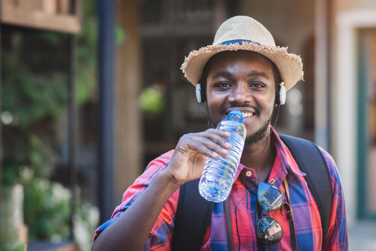 A smiling man drinking bottled water while outdoors, wearing a backpack, hat, headphones, and sunglasses.