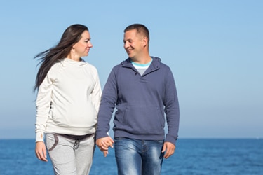 A pregnant woman holds hands with her partner while walking by the water.